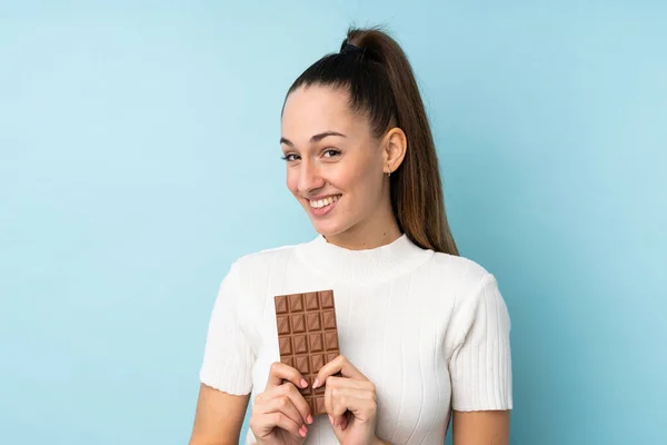 Young Brunette Woman Isolated Blue Background Taking Chocolate Tablet Happy — Stock Photo, Image