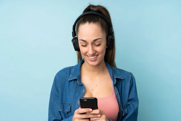 Young Brunette Woman Isolated Blue Background Listening Music Looking Mobile — Stock Photo, Image