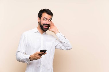 Sakallı genç adam bir cep telefonu tutuyor. Kulağına elini sürerek bir şeyler dinliyor.