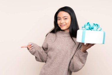 Genç Asyalı kız elinde büyük bir pasta tutuyor. Bej arka planda izole edilmiş. Şaşırmış ve parmağıyla işaret ediyor.