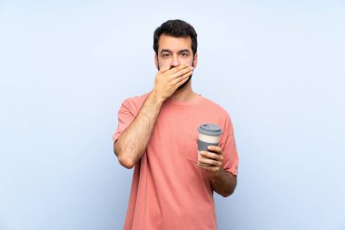 Sakallı genç adam, izole edilmiş mavi arka planda elleri ve ağzı kapalı bir kahve tutuyor.