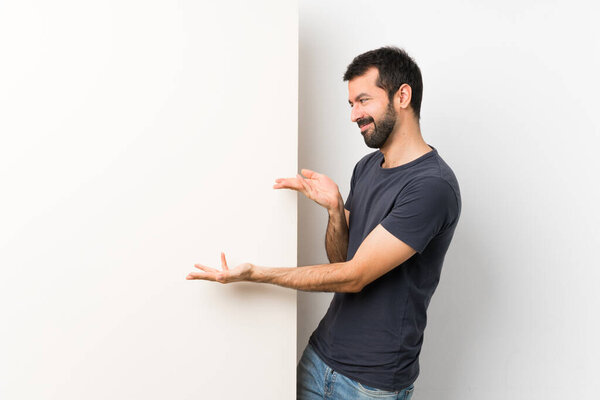 Young handsome man with beard holding a big empty placard extending hands to the side for inviting to come