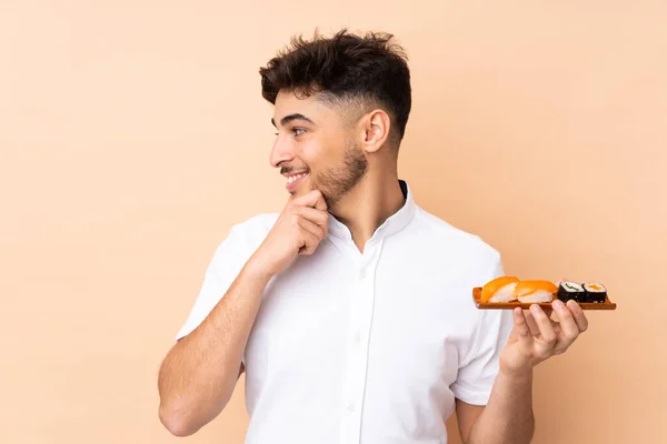 Arabian man eating sushi isolated on beige background thinking an idea and looking side