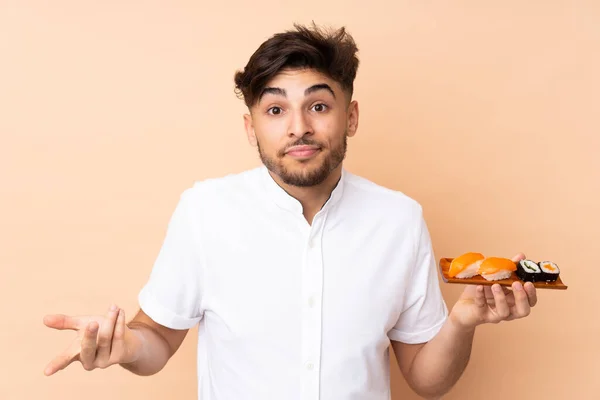 Arabian man eating sushi isolated on beige background making doubts gesture while lifting the shoulders