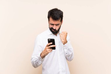 Sakallı genç adam elinde kızgın bir hareketle cep telefonu tutuyor.