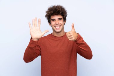 İzole edilmiş mavi arka planda genç beyaz adam parmakları olan altı kişi sayıyor.