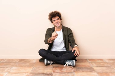 Genç, beyaz bir adam yerde oturuyor ve parmağını kaldırıyor.