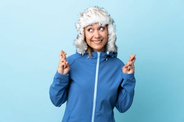 Kış şapkalı genç Uruguaylı kız mavi arka planda izole edilmiş ve şans diliyor.