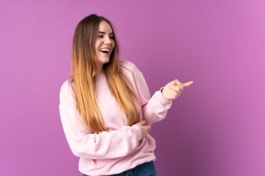 Mor arka planda izole edilmiş genç beyaz bir kadın parmak göstererek bir ürün sunuyor.