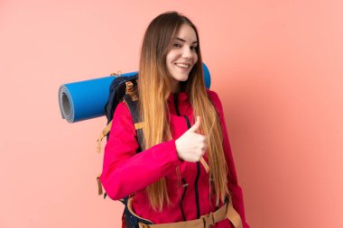 Pembe arka planda izole edilmiş büyük sırt çantalı genç dağcı kız baş parmak işareti yapıyor.