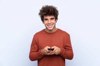 İzole edilmiş mavi arka plan üzerinde genç beyaz adam cep telefonuyla bir mesaj gönderiyor.