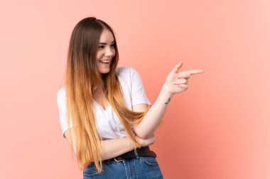 Pembe arka planda izole edilmiş genç beyaz bir kadın parmak göstererek bir ürün sunuyor.