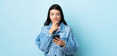 Genç esmer kız izole edilmiş mavi arka planda düşünüyor ve mesaj gönderiyor.