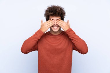 İzole edilmiş mavi arka planda genç beyaz adam elleriyle gözlerini kapatıyor.