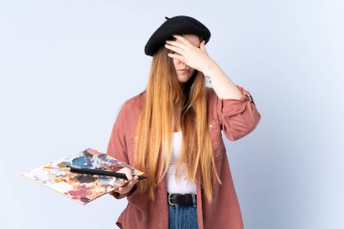 Mavi arka planda bir palet tutan genç sanatçı kadın gözlerini elleriyle kapatıyor.