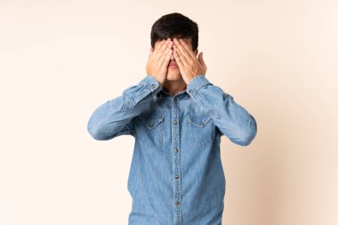 İzole edilmiş bej arka planda yakışıklı bir adam. Gözlerini elleriyle kapatıyor.