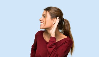 Sarışın kadın izole mavi arka plan üzerinde kulak elini koyarak bir şey dinlerken