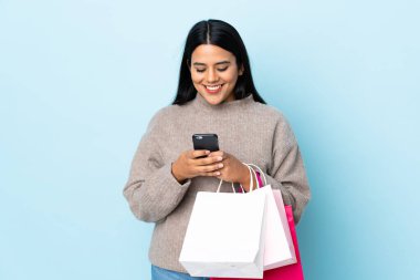 Genç Latin bir kadın mavi arka planda izole edilmiş. Elinde alışveriş poşetleri var ve bir arkadaşına cep telefonuyla mesaj yazıyor.