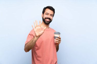 Elinde kahve tutan sakallı genç adam izole edilmiş mavi arka planda beş parmaklı sayıyor.