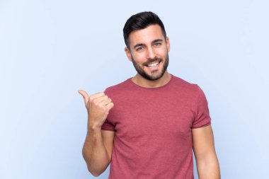İzole edilmiş mavi arka planı olan sakallı yakışıklı genç bir adam bir ürün sunmak için kenarı işaret ediyor.