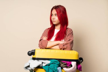 Bir bavul dolusu elbisesi olan gezgin kız bej arka planda izole edilmiş kollarını çapraz tutuyor.