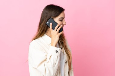 Pembe arka planda izole edilmiş genç beyaz bir kadın cep telefonuyla sohbet ediyor.