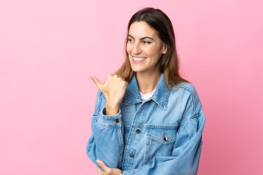 Pembe arka planda izole edilmiş genç beyaz kadın bir ürünü sunmak için kenarı işaret ediyor.