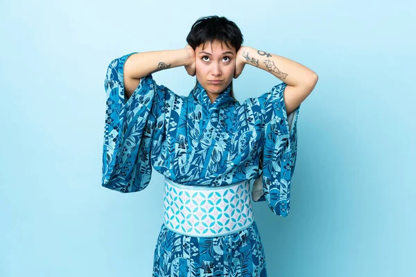 İzole edilmiş mavi arka planda kimono giyen genç bir kadın hayal kırıklığına uğramış ve kulaklarını kapatmış.