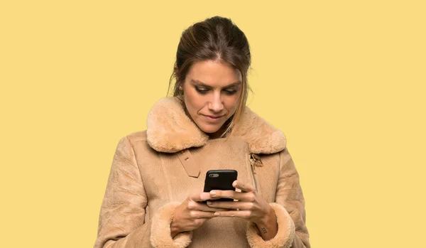 Blondýnka Kabátem Který Posílá Vzkaz Mobilními Přes Izolované Žluté Pozadí — Stock fotografie