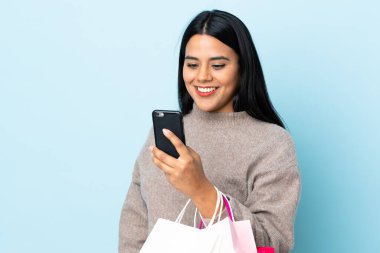 Genç Latin bir kadın mavi arka planda izole edilmiş. Elinde alışveriş poşetleri var ve bir arkadaşına cep telefonuyla mesaj yazıyor.