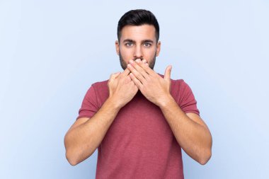 Sakallı, genç ve yakışıklı bir adam, izole edilmiş mavi arka planda, ağzı elleriyle kapatıyor.