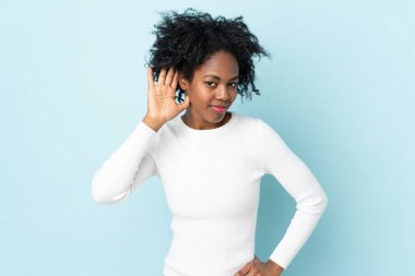 Genç Afrikalı Amerikalı bir kadın mavi arka planda izole edilmiş. Kulağına elini sürerek bir şeyler dinliyor.