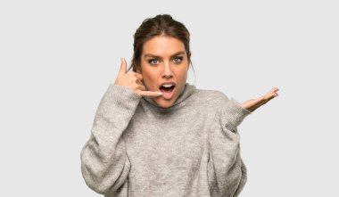 Balıkçı yakalı sarışın kadın telefon hareketi yapıyor ve izole edilmiş gri arka plandan şüphe ediyor.