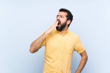 Sakallı genç adam izole edilmiş mavi arka planda esniyor ve ağzını eliyle açık tutuyor.