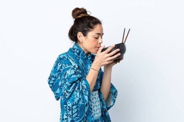 İzole edilmiş beyaz arka planda kimono giyen bir kadın elinde yemek çubuklarıyla bir kase erişte tutuyor ve onu yiyor.