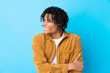 Kadife ceketli genç Afro-Amerikalı adam mavi arka planda gülüyor.