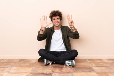 Genç beyaz adam yerde oturmuş parmaklarıyla sekizi sayıyor.