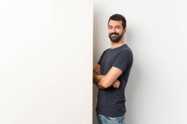 Sakallı genç yakışıklı bir adam. Elinde kocaman boş bir pankart var. Kolları çapraz ve ileriye bakıyor.