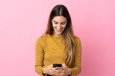 Pembe arka planda izole edilmiş genç beyaz kadın cep telefonuyla bir mesaj gönderiyor.