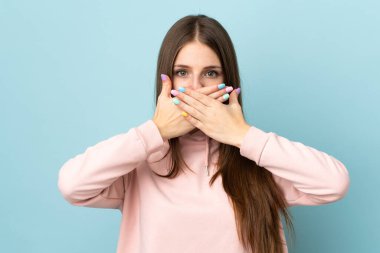 Mavi arka planda izole edilmiş genç beyaz kadın elleriyle ağzını kapatıyor.