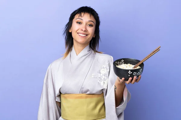 Young woman wearing kimono over isolated blue background smiling a lot while holding a bowl of noodles with chopsticks