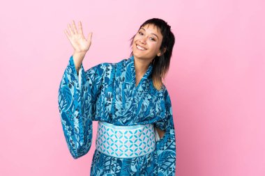 İzole edilmiş mavi arkaplanda kimono giyen genç bir kadın mutlu bir ifadeyle selamlıyor.