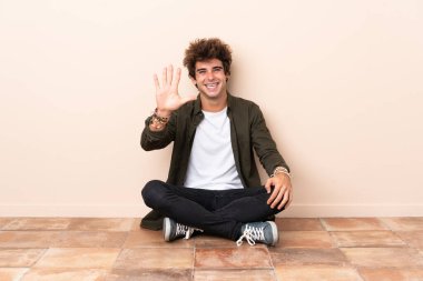 Beyaz bir adam yerde oturmuş parmaklarıyla beşi sayıyor.