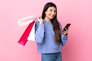 Soyutlanmış pembe arka planda genç esmer kız elinde alışveriş poşetleri ve cep telefonuyla bir arkadaşına mesaj yazıyor.