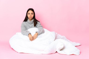 Genç melez kadın pijama giymiş yerde üzgün bir ifadeyle oturuyor.
