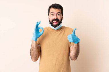 Koronavirüsten sakallı, maskeli ve eldivenli beyaz bir adam izole bir arkaplanda işaret veriyor.