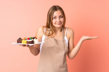 Pastacı Ukraynalı şef pembe arka planda izole edilmiş bir kek tutuyor. Kafası karışık bir yüz ifadesi var.