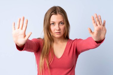 Genç Ukraynalı kız mavi arka planda izole edilmiş. Durma hareketi yapıyor ve hayal kırıklığına uğruyor.