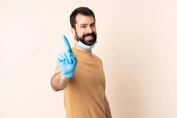 一个白种人 留着胡子 戴着面具和手套 戴在孤立的背景上 露出并举起一只手指 保护自己不受头皮的侵害 — 图库照片