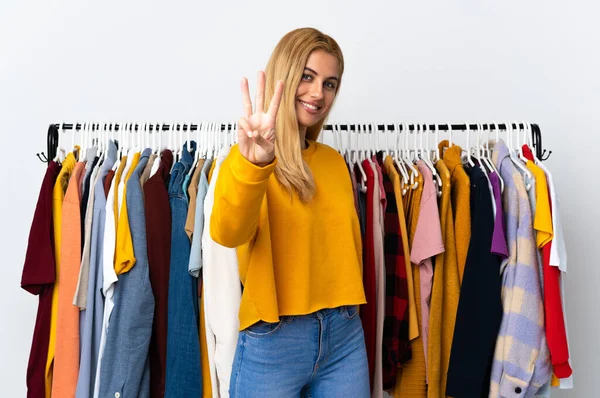 Young Uruguayan blonde woman in a clothing store happy and counting three with fingers